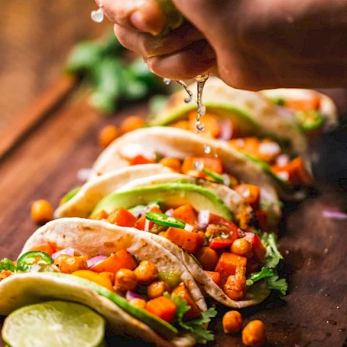 A person sprinkles toppings onto tacos with fresh veggies and lime on a wooden surface.