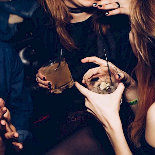 A party scene where a woman holds two drinks with straws, surrounded by friends with dark, glam makeup and jewelry, enjoying the night.