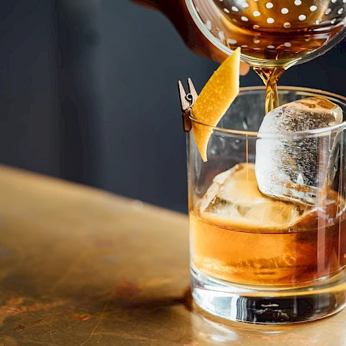 An amber cocktail is being poured into a rocks glass over ice, with a citrus garnish hooked on the rim.