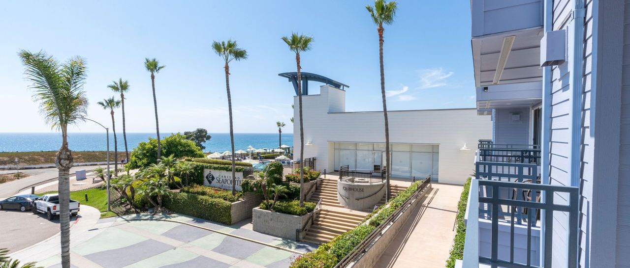 A sunny seaside resort courtyard with palm trees, a walkway, and an ocean view beside a white building, overlooking the coast.