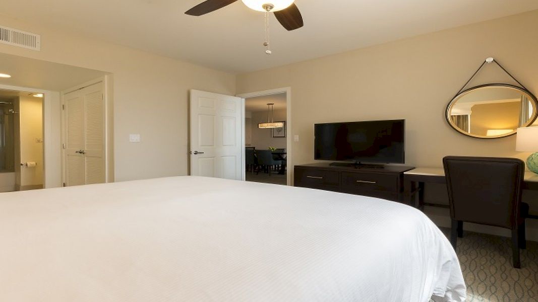 A clean hotel room with a large bed in the foreground, a desk and chair, a wall-mounted TV, a round mirror, and a doorway leading to a bathroom area.