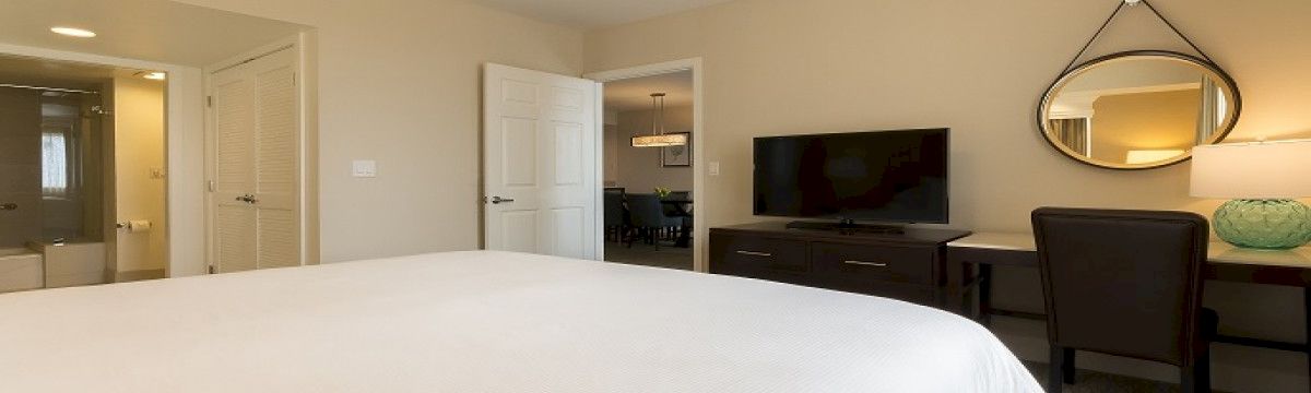 A clean hotel room with a large bed in the foreground, a desk and chair, a wall-mounted TV, a round mirror, and a doorway leading to a bathroom area.