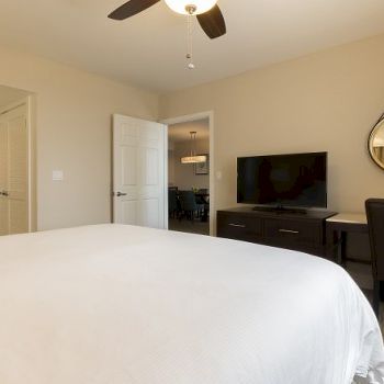 A tidy hotel bedroom with a large white bed, wall mirror, floating desk with two chairs, TV console, ceiling fan, and soft beige walls.