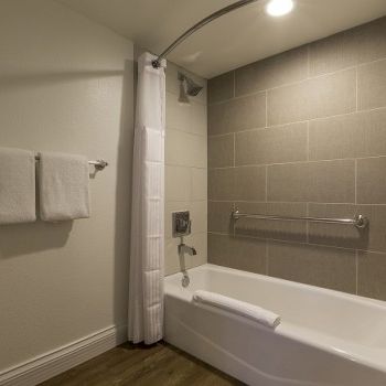 A modern bathroom with a white bathtub, gray tiled wall, shower curtain, towel rack with two white towels, and a dark wood floor. 140 chars total.