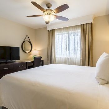 A tidy hotel room with a large bed, bedside lamps, a ceiling fan, a desk with a TV, and a window with curtains.