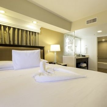 A tidy hotel room with a large bed, white linens, decorative headboard, bedside lamp, and a doorway to a small suite area with a sink and mirror.