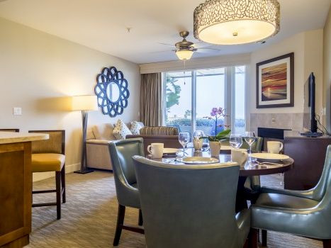 A cozy living area with a round dining table, four teal chairs, another sofa, a wall mirror, and a balcony view in the background.