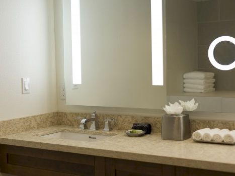 A modern bathroom vanity with a granite counter, sink, stacked towels, a tissue box, and a circular mirror with built-in light on the right.