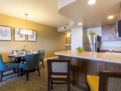 A modern apartment kitchen and dining area with a breakfast bar, teal chairs, a round dining table set for four, warm yellow walls, and stainless appliances.