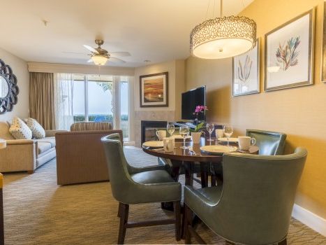 A modern hotel suite with a dining area, circular table and green chairs, a living room in the background, warm lighting, and decor.