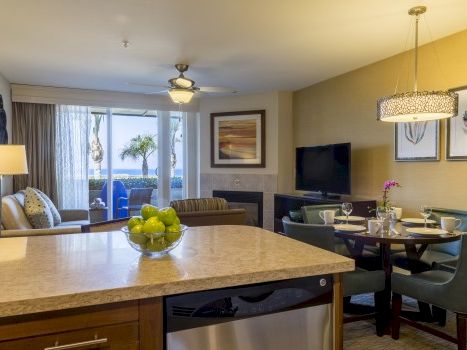 A cozy, modern living area with a kitchen island, seating for six, a wall-mounted TV, plants, framed art, and a sliding door to a balcony.