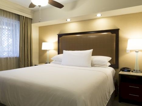 A clean hotel room with a king bed, white linens, a padded headboard, two bedside tables with lamps, and a ceiling fan above the bed.