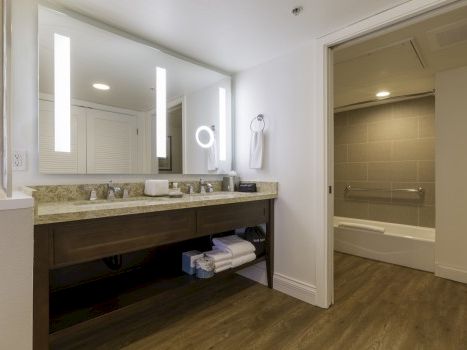 A modern bathroom with a double vanity, large backlit mirror, dark wood cabinet, marble countertop, and a separate tiled shower/tub area.