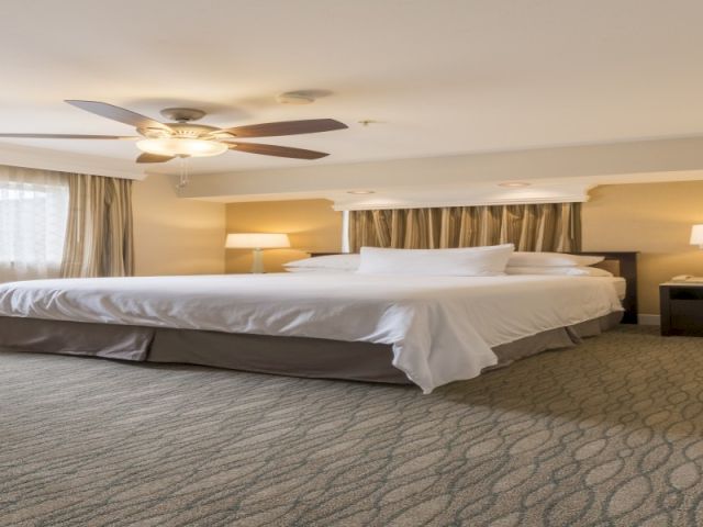 A spacious hotel room with a large bed, two nightstands, ceiling fan, soft lighting, and a patterned carpet.