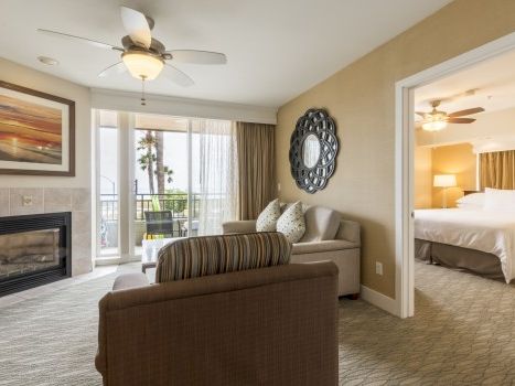 A modern hotel suite: living area with couch, fireplace, wall art, ceiling fan, balcony view, and a connected bedroom with a king bed, soft lighting, and neutral decor.
