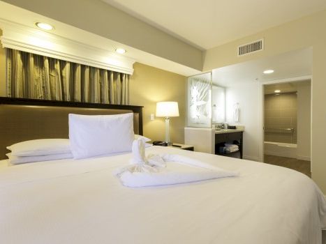 A tidy hotel bedroom with a large bed, white linens, decorative headboard, bedside lamp, a neat towel sculpture, and a bright adjoining bathroom.