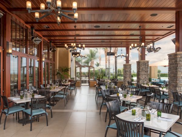 Covered outdoor dining patio with warm lights, stone pillars, and set tables and chairs, with palm trees outside.