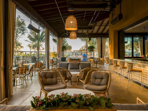Sunlit hotel patio with wicker lounge chairs, bar seating, hanging pendant lanterns, and a view of palm trees at dusk.