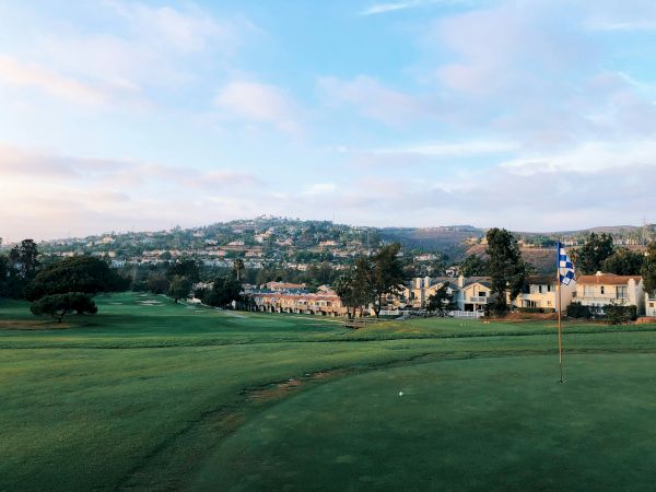 A green golf course with a flag on the green, rolling hills, and a cluster of buildings in the distance under a partly cloudy sky.