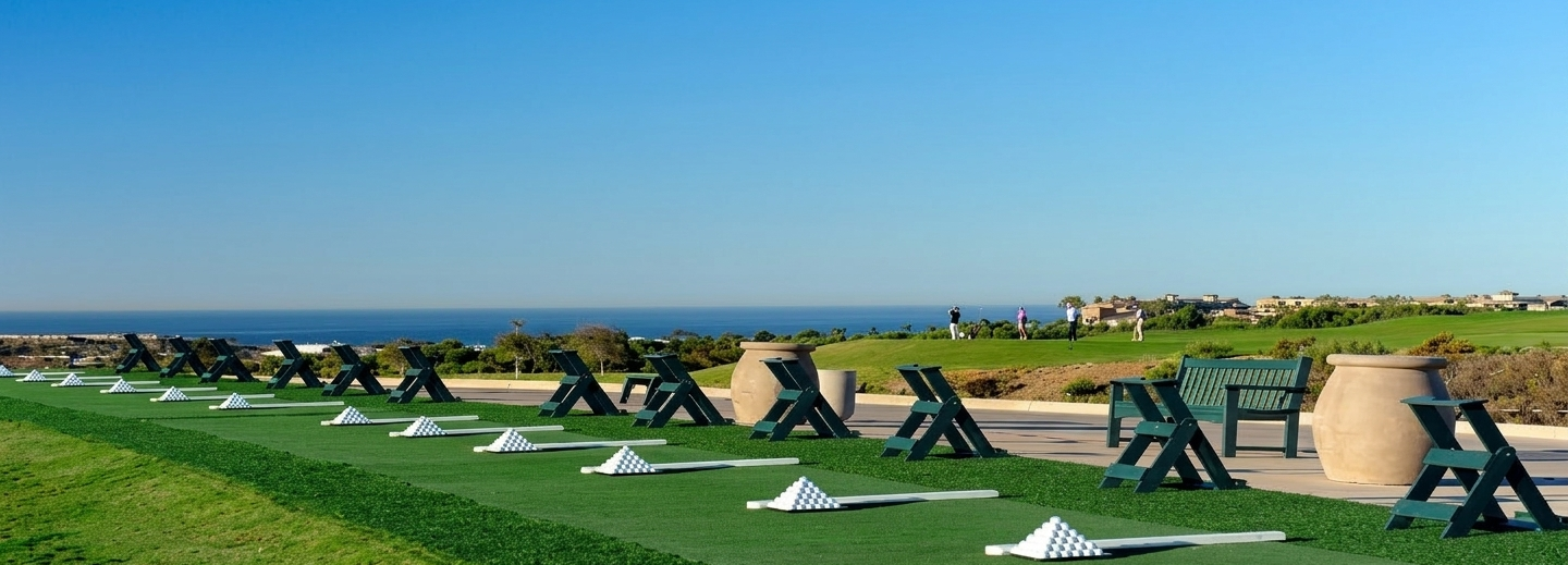 A coastal driving range with neatly arranged black-green practice stands, chequered golf mats, and lush green turf under a bright blue sky.