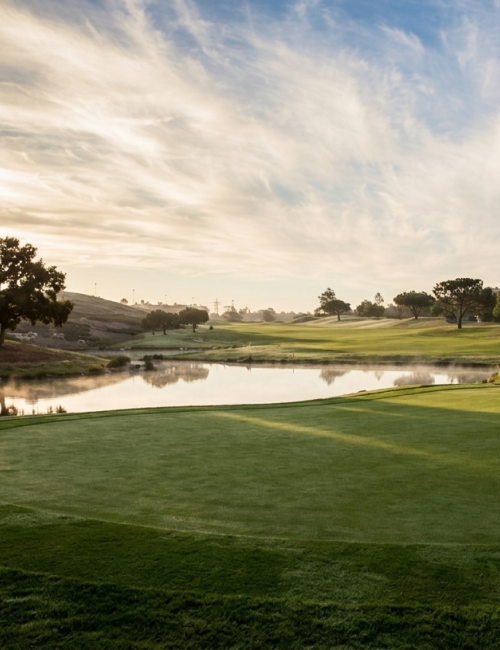 A serene golf course at sunset with a calm water hazard, green fairways, scattered trees, and a distant cluster of houses under a dramatic sky.
