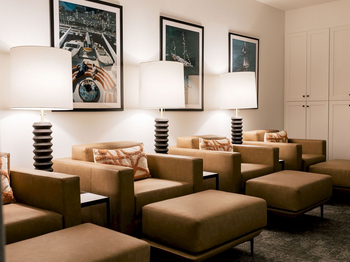 A modern hotel lobby seating area with row of brown armchairs, ottomans, lamps, framed photos, and a white closet wall in the background.