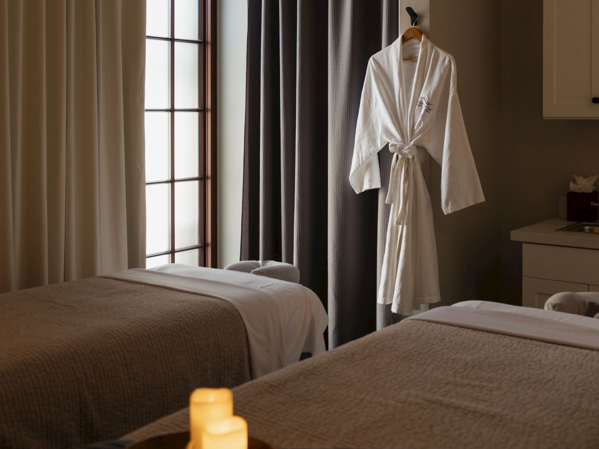 A cozy hotel bedroom with two beds, dark and light curtains by a window, a white bathrobe on a hanger, and a lit candle tray.