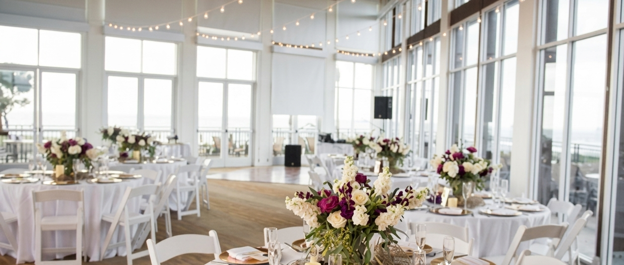 Guests dine in a bright, elegant reception hall with white linens, florals, and string lights, set for a formal wedding or event.