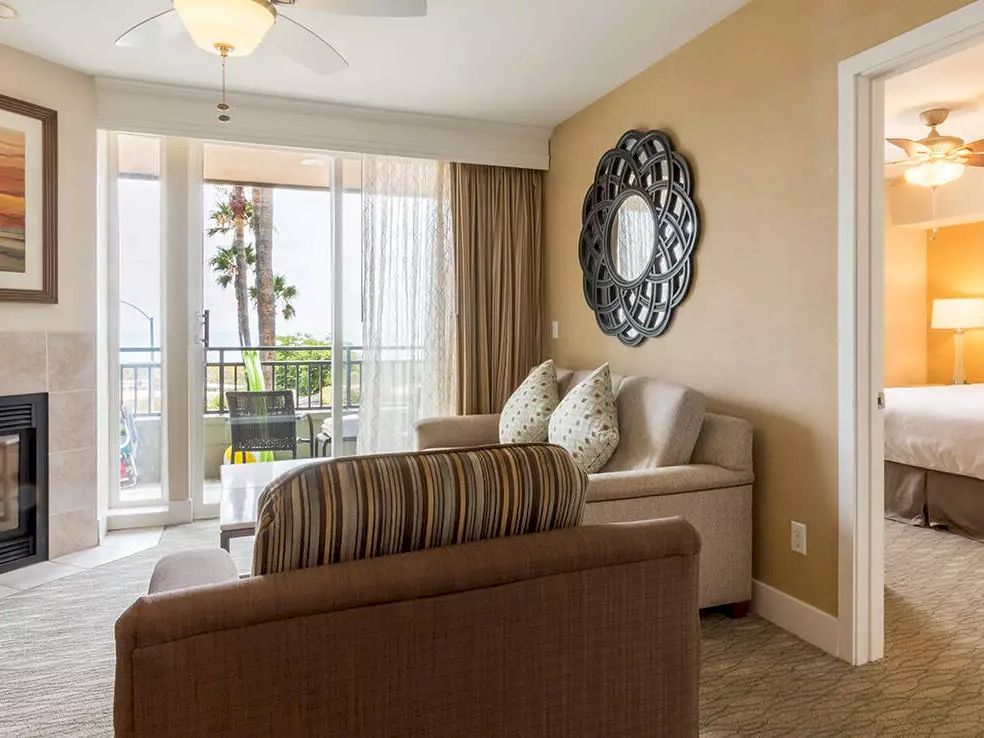 A cozy hotel suite with a living area, fireplace, balcony, and a connected bedroom with a king bed, neutral decor, and soft lighting.