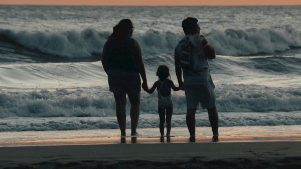 A family of three holds hands at the shore during a sunset, silhouettes against the waves as the light fades.