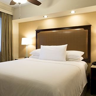 A cozy hotel bedroom with a large bed, warm lighting, marble-topped nightstands, and a window with curtains.