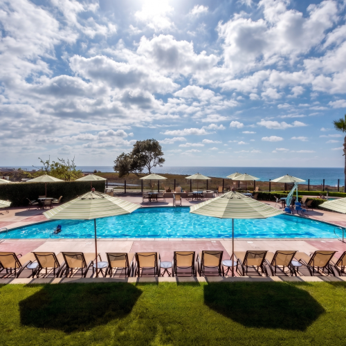 Sunny resort pool with striped umbrellas, lounge chairs, tall palm trees, and the ocean under dramatic clouds.