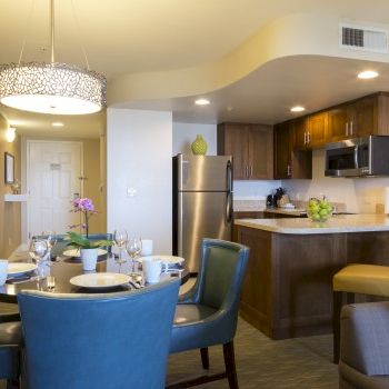A modern open-plan dining and kitchen area with brown wood cabinets, stainless fridge, round dining table, blue chairs, decorative lighting, and a small lounge corner.