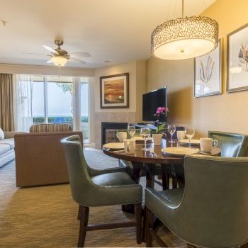Bright dining/living room with a glass-top table, green chairs, ceiling light, and framed wall art.