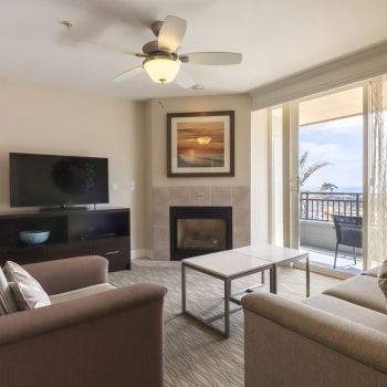 A cozy living room with a wall TV, fireplace, beige sofas, a coffee table, and a balcony with ocean views through glass doors, ending this sentence.