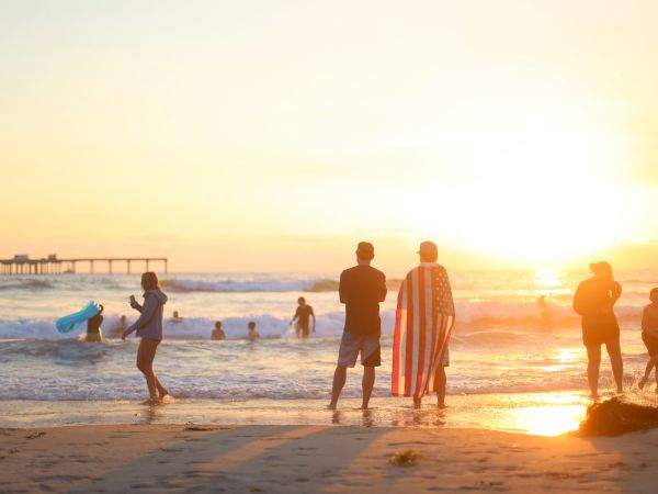 People enjoy a beach sunset, with a man carrying an American flag as waves glow in the golden light.